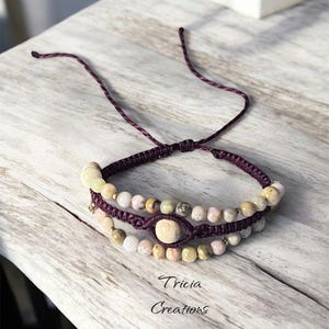 Peut inclure: Un bracelet macramé fait à la main avec trois rangées de perles en pierre naturelle dans les tons crème, rose et beige. Le bracelet est fixé avec un cordon violet foncé et porte le texte "Tricia Creations".
