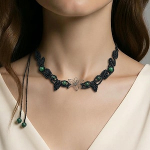 May include: A black macrame necklace featuring a central silver-colored flower pendant and interspersed green beads. The necklace has a leaf-like design and adjustable ties with matching green beads.