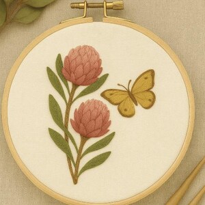 May include: Embroidered floral design with two pink flowers, green leaves, and a yellow butterfly, set in a wooden embroidery hoop. The text "Sarah & Bee Designs" is visible. The design is on cream-coloured fabric. The hoop is approximately 20 cm in diameter.