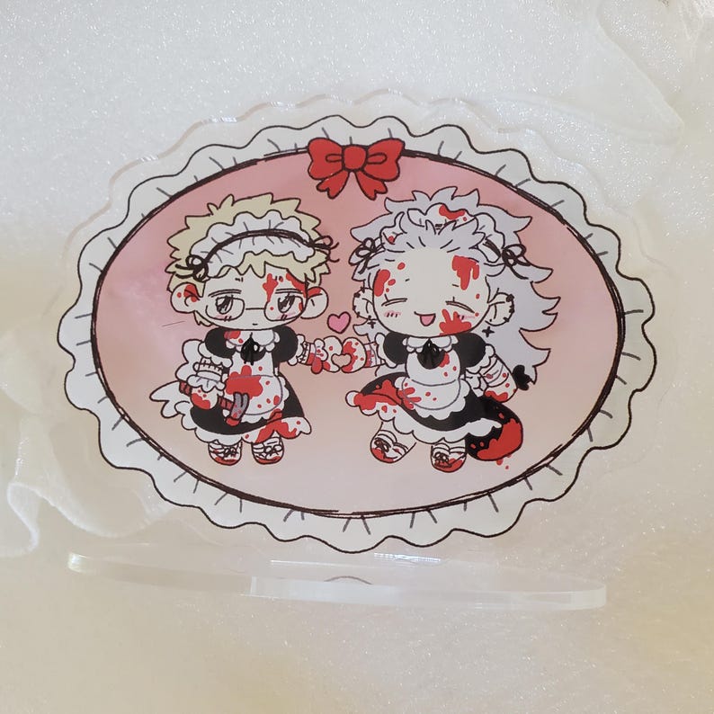 May include: A pink and white oval-shaped acrylic stand with a black lace border. The stand features a cartoon illustration of two characters dressed in maid outfits with blood splatters. The characters are holding hands and smiling.