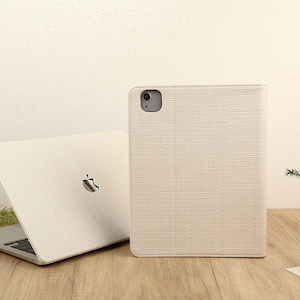 May include: A white laptop computer with a white case and a white keyboard. The laptop is closed and sitting on a wooden table. Next to the laptop is a white case for a tablet or phone. The case has a textured fabric design.