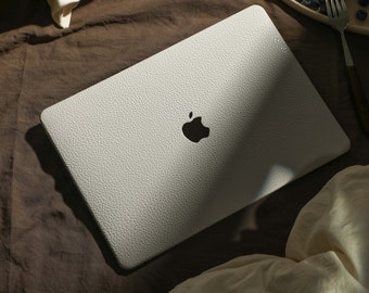 May include: A white laptop computer with a textured surface and an Apple logo on the lid. The laptop is on a brown fabric surface with a white pillow and a plate of blueberries and a bagel with cream cheese.