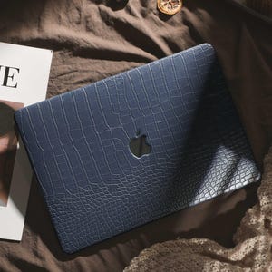 May include: A navy blue laptop case with a crocodile skin texture. The case has a cutout for the Apple logo. The laptop case is on a brown surface with a magazine and a beige knitted blanket.