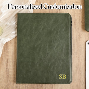 Puede incluir: Una funda para iPad de cuero verde oscuro con un monograma dorado "SB" en la parte delantera. La funda está sobre una superficie de madera con una tela blanca y una tableta blanca.