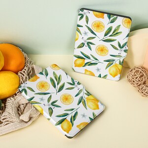 May include: Two white e-reader cases with a lemon and leaf pattern. The cases are on a light yellow surface with lemons and a woven basket.