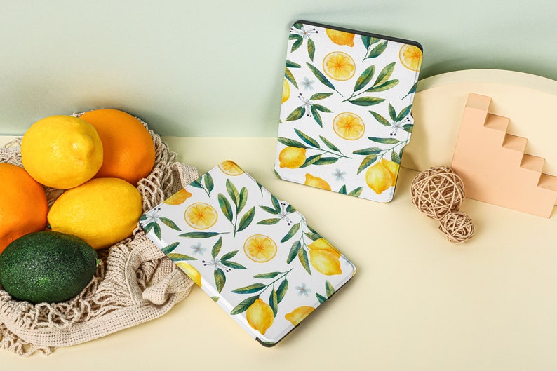 May include: Two white e-reader cases with a lemon and leaf pattern. The cases are on a light yellow surface with lemons and a woven basket.