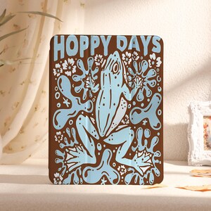 Puede incluir: Una funda ilustrada marrón y azul con una rana y el texto "Hoppy Days".