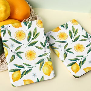 May include: Two white e-reader cases with a lemon and leaf pattern. The cases are on a light yellow surface with lemons and a green avocado in the background.
