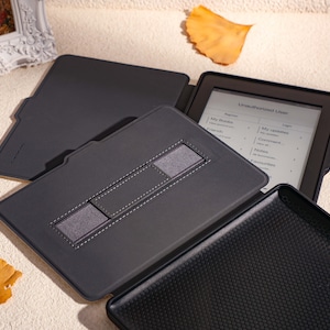 May include: A black e-reader case with a built-in stand and a strap to hold the e-reader in place. The e-reader is open and displaying a menu with the text "Library", "My Books", "My Collections", "Login", "Recent", "Settings", "Help", and "Favorites".