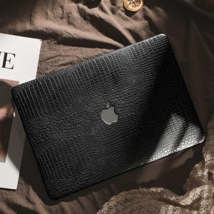May include: Black laptop case with a crocodile skin effect. The case has a silver Apple logo. The laptop case is on a brown surface. The case has a textured surface.