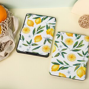 May include: Two white e-reader cases with a lemon and leaf pattern. The cases are on a light yellow surface.