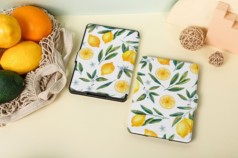 May include: Two white e-reader cases with a lemon and leaf pattern. The cases are on a light yellow surface.