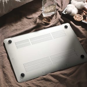 May include: A silver laptop computer with black rubber feet rests on a brown fabric. The underside of the laptop features ventilation slots. A glass bottle, cotton, and dried citrus slices are also visible.