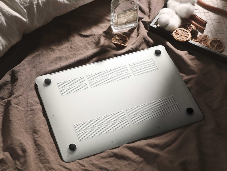 May include: A silver laptop computer with black rubber feet rests on a brown fabric. The underside of the laptop features ventilation slots. A glass bottle, cotton, and dried citrus slices are also visible.