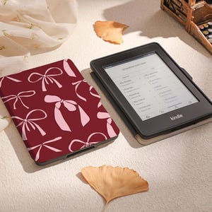 May include: A burgundy Kindle e-reader case with a pink bow pattern. The case is lying on a white surface with a white and pink fabric in the background.