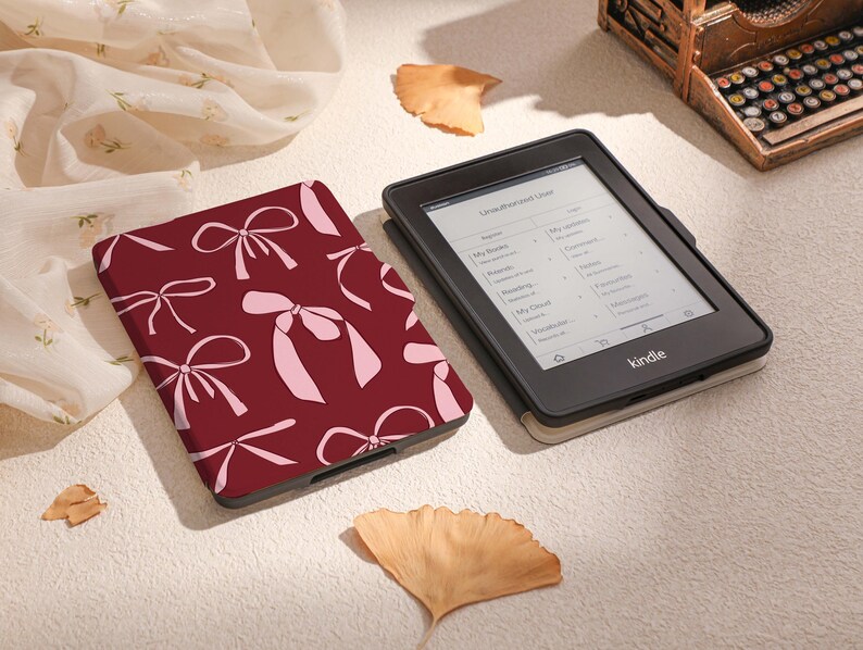May include: A burgundy Kindle e-reader case with a pink bow pattern. The case is lying on a white surface with a white and pink fabric in the background.