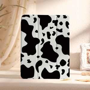 May include: A tablet case featuring a cow print design. The case has a white background with irregular black spots. The folding cover is visible. The case is on a beige surface.