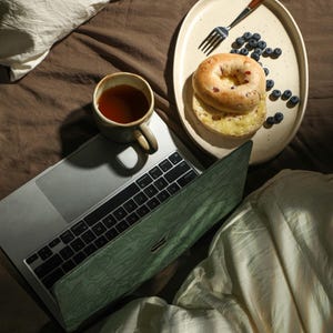 May include: A laptop computer with a green cover is on a bed with a white blanket. A plate with a bagel, blueberries, and a fork is on the bed. A cup of tea is on the laptop.