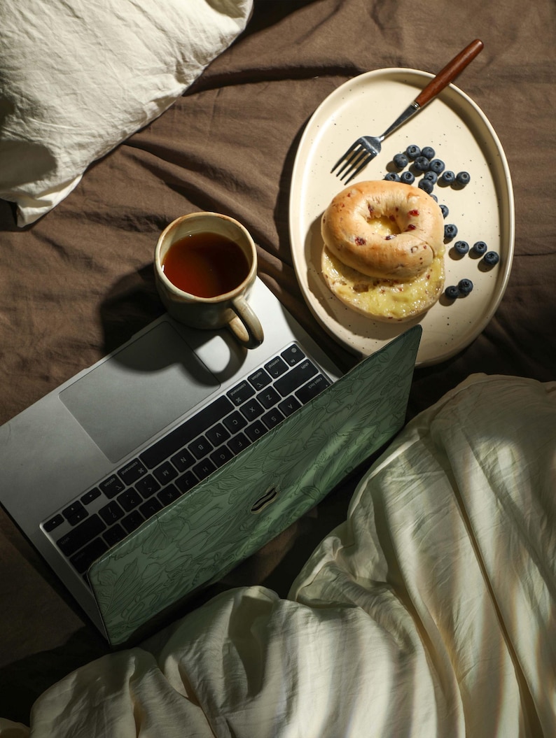 May include: A laptop computer with a green cover is on a bed with a white blanket. A plate with a bagel, blueberries, and a fork is on the bed. A cup of tea is on the laptop.