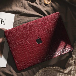 Puede incluir: Una funda para portátil de color burdeos con un patrón texturizado similar a la piel de cocodrilo. La funda presenta un recorte del logotipo de Apple en negro. La funda está sobre una superficie marrón con una revista y otros objetos en el fondo.