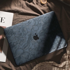 Peut inclure: Une coque d'ordinateur portable bleu foncé avec un motif floral en relief. L'étui présente un motif détaillé de fleurs et de feuilles, avec le logo Apple visible. L'étui pour ordinateur portable est sur une surface marron.