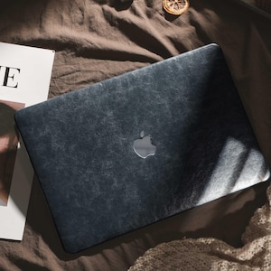 May include: A dark blue laptop with a silver Apple logo on a brown surface. The laptop has a textured, leather-like appearance. A magazine with the letter "E" visible is next to the laptop.