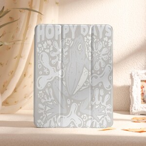 Puede incluir: Funda blanca para iPad con una ilustración de rana y el texto "Hoppy Days".
