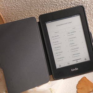 May include: A black e-reader with a screen displaying an "Unauthorized User" message. The screen shows options to register or log in, and lists various features such as "My Books", "Friends", "Reading", "My Cloud", "Vocabular", "My updates", "Comment", "Notes", "Favourites", and "Messages". The e-reader is open and has a black cover.