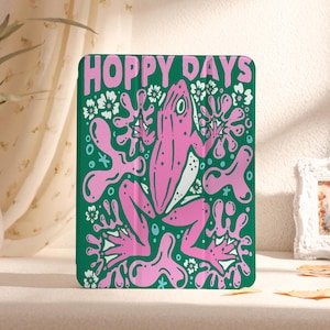 Puede incluir: Una funda verde para iPad con una ilustración de rana rosa y el texto "Hoppy Days". La rana está rodeada de diseños florales rosas y blancos.