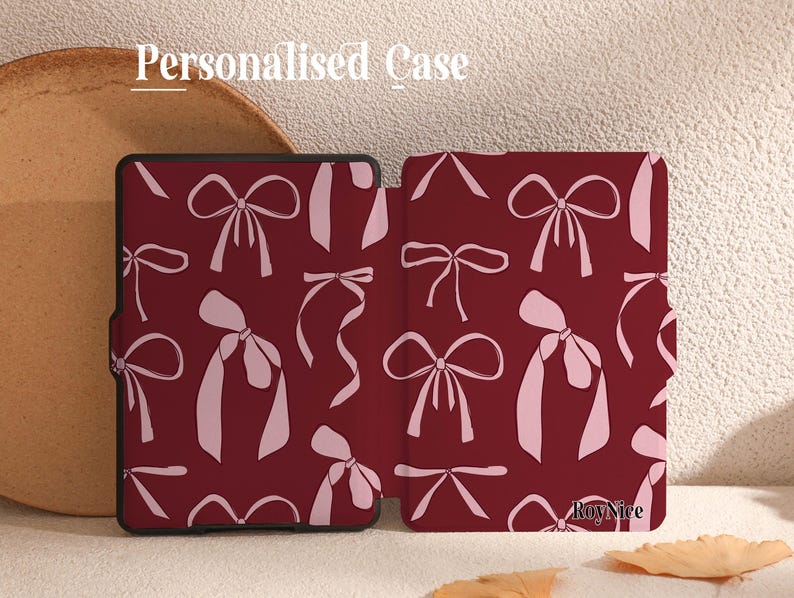 May include: A personalised case for an e-reader with a burgundy background and a pattern of pink bows. The case has the text "Roy Nice" printed on the bottom right corner.