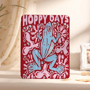 Puede incluir: Funda de teléfono de color rojo y rosa con el texto "Hoppy Days" y una ilustración de una rana azul.