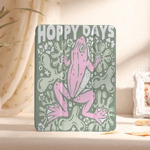 Puede incluir: Una funda para iPad verde y rosa con una rana y el texto "Hoppy Days".
