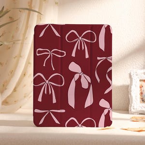 May include: A burgundy iPad case with a pink bow pattern. The case features a variety of bow designs, including a large bow in the centre.