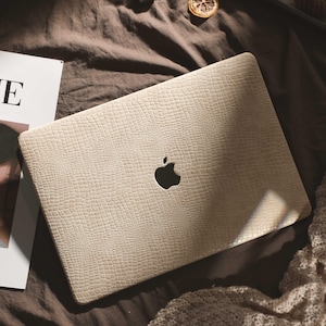 May include: A beige laptop case with a reptile skin effect. The case features a cut-out for the Apple logo. The laptop is on a brown surface, with a magazine and other decorative items nearby.