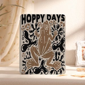 Puede incluir: Una ilustración de rana marrón y negra con el texto "Hoppy Days" sobre un fondo blanco. La rana está rodeada de formas abstractas y flores negras y blancas.