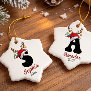 Könnte beinhalten: Zwei weiße, schneeflockenförmige Keramikornamente mit goldenen Aufhängeschnüren. Eines zeigt den schwarzen Buchstaben S mit Rentiergeweih und Weihnachtsmütze, den Namen Sophia und 2026. Das andere zeigt den Buchstaben A, den Namen Amadar und 2024.
