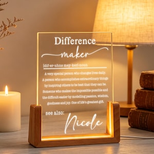 Könnte beinhalten: Ein beleuchtetes Acrylschild mit dem Text "Difference maker" und einer Definition. Das Schild befindet sich in einem Holzrahmen und steht auf einem Tisch. Eine Kerze, eine Lampe und Bücher sind im Hintergrund.