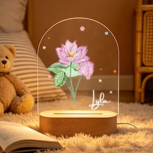 Könnte beinhalten: Ein dekoratives Nachtlicht mit einem Design einer rosa Lotusblume, grünen Blättern und dem Namen "Lyla" in Schreibschrift. Das Licht steht auf einem Holzsockel und wird über ein Kabel mit Strom versorgt.