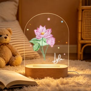 May include: A decorative night light with a pink lotus flower design, green leaves, and the name "Lyla" in cursive. The light sits on a wooden base and is powered by a cord.