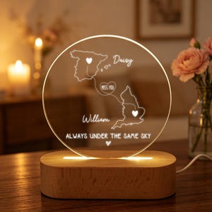 May include: A personalized night light with a wooden base and a clear, circular acrylic top. The acrylic features illuminated outlines of Spain and England, with names and the phrase "ALWAYS UNDER THE SAME SKY". The light emits a warm glow.