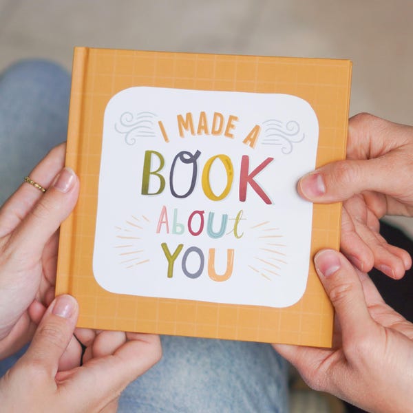 May include: A small, square book with an orange cover and a white rectangular design. The design features the text "I MADE A BOOK ABOUT YOU" in colorful, playful lettering. The book is held by two sets of hands.