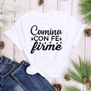 Puede incluir: Camiseta blanca con la frase "Camina con fe firme" en escritura negra. La camiseta est&aacute; anudada a la cintura y combinada con unos vaqueros azules. Pi&ntilde;as y vegetaci&oacute;n se disponen alrededor de la camiseta sobre un fondo de madera clara.