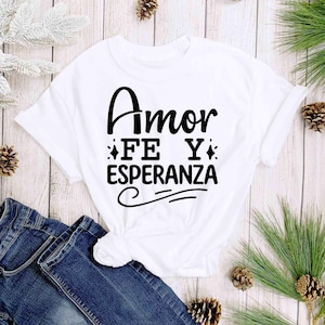 Puede incluir: Camiseta blanca con las palabras "Amor Fe Y Esperanza" en escritura negra. La camiseta est&aacute; anudada a la cintura y combinada con unos vaqueros azules. Agujas de pino y pi&ntilde;as decoran el fondo.