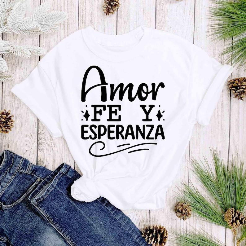 Puede incluir: Camiseta blanca con las palabras "Amor Fe Y Esperanza" en escritura negra. La camiseta est&aacute; anudada a la cintura y combinada con unos vaqueros azules. Agujas de pino y pi&ntilde;as decoran el fondo.