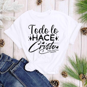 Puede incluir: Camiseta blanca con el texto en espa&ntilde;ol "Todo lo HACE Cristo" en escritura negra. La camiseta est&aacute; anudada a la cintura y se muestra con vaqueros azules. El fondo incluye agujas de pino y pi&ntilde;as.
