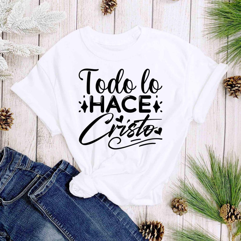 Puede incluir: Camiseta blanca con el texto en espa&ntilde;ol "Todo lo HACE Cristo" en escritura negra. La camiseta est&aacute; anudada a la cintura y se muestra con vaqueros azules. El fondo incluye agujas de pino y pi&ntilde;as.