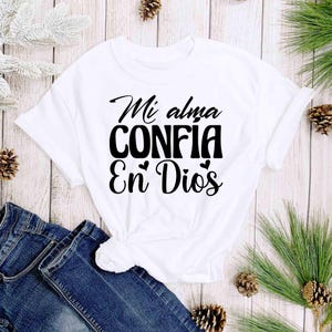 Puede incluir: Camiseta blanca con la frase en espa&ntilde;ol "Mi alma CONFIA En Dios" en escritura negra. La camiseta est&aacute; anudada a la cintura y se combina con unos vaqueros azules. El fondo incluye agujas de pino y pi&ntilde;as.
