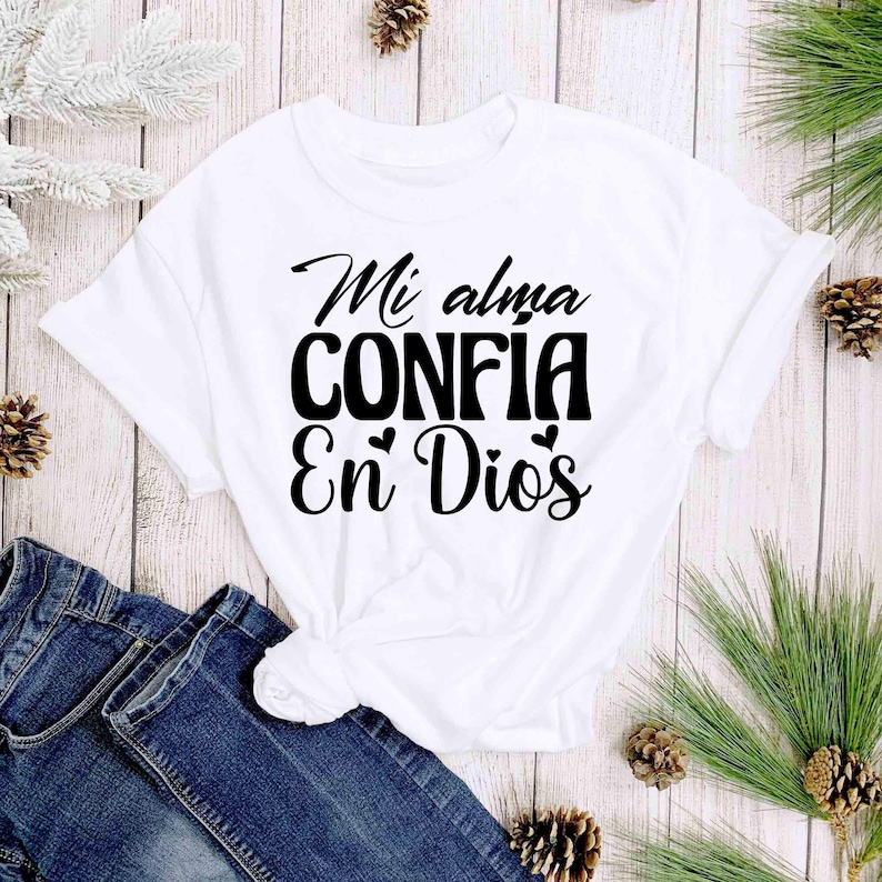 Puede incluir: Camiseta blanca con la frase en espa&ntilde;ol "Mi alma CONFIA En Dios" en escritura negra. La camiseta est&aacute; anudada a la cintura y se combina con unos vaqueros azules. El fondo incluye agujas de pino y pi&ntilde;as.