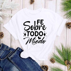 Puede incluir: Camiseta blanca con la frase en espa&ntilde;ol "FE Sobre TODO Miedo" en escritura negra. La camiseta est&aacute; anudada a la cintura y combinada con unos vaqueros azules. El fondo incluye agujas de pino y pi&ntilde;as.