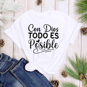 Puede incluir: Camiseta blanca con la frase "Con Dios TODO ES Posible" en escritura negra. La camiseta se combina con vaqueros azules y est&aacute; decorada con pi&ntilde;as y vegetaci&oacute;n.